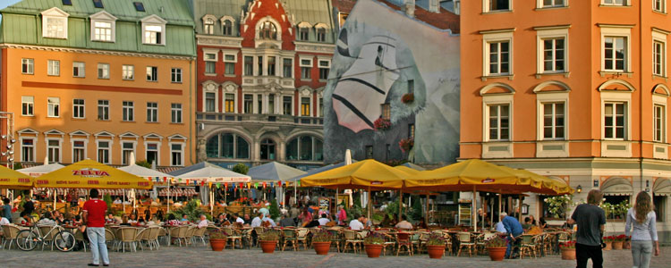 People eating and relaxing in Riga’s Dome Square. One of the buildings features a mural depicting a glider plane preparing for takeoff. The spire of St. Peter’s Church is visible in the background.