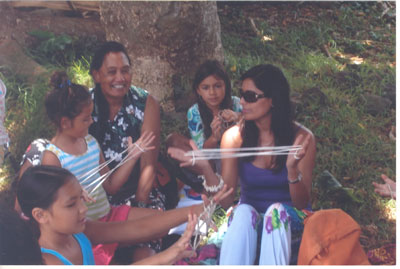A group lesson on kai-kai, one of the oldest traditional Rapa Nui skills, consisting of telling a story through figures made on a loop of string. A teacher, Isabel, is sitting behind the children and a friend. 