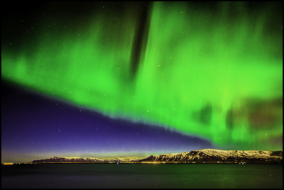 Dayglow-green lights over a dark-blue horizon and tan mountains, all reflected in water — Iceland, February 2014. Photo by Nelson Burack