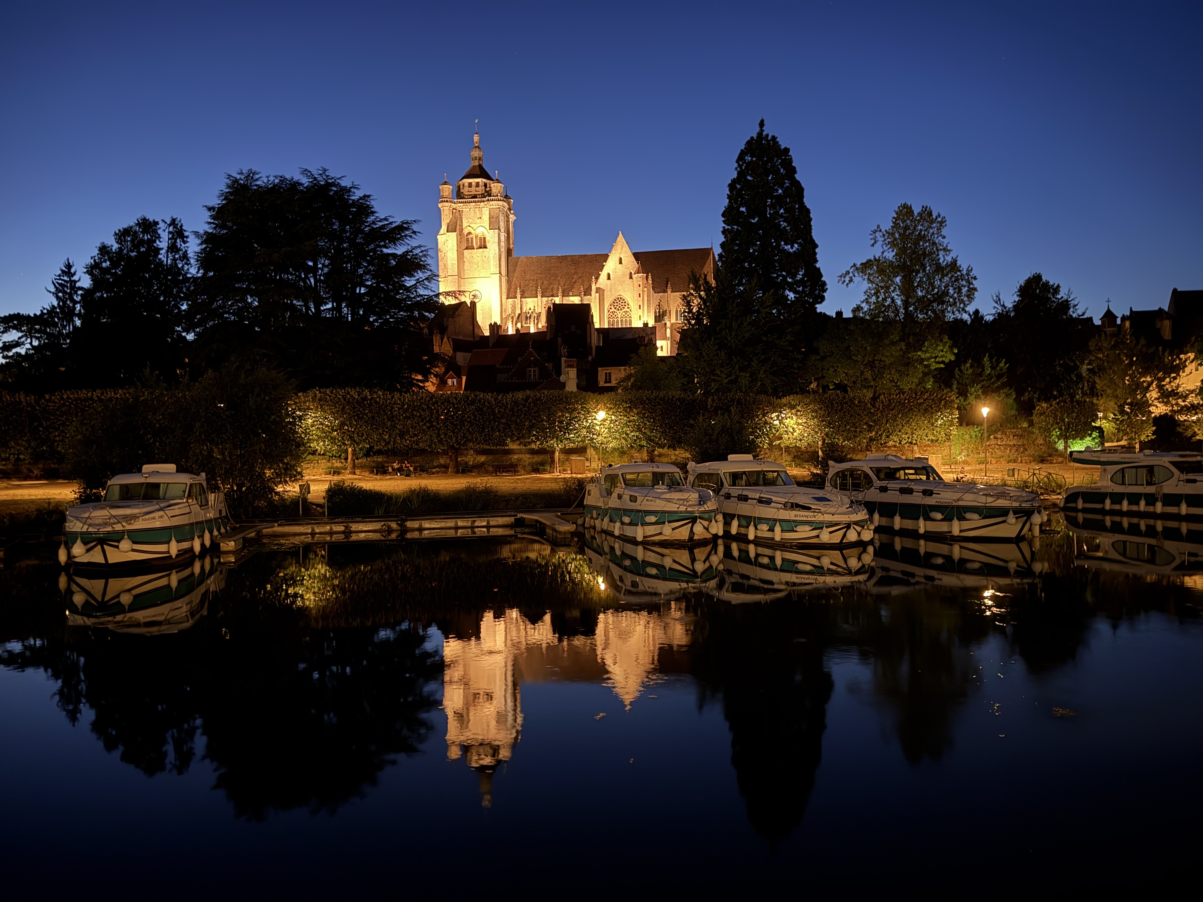 The Collégiale Notre-Dame de Dole glows above the canal at dusk in Dole, France, reflected in the still water alongside moored canal boats.