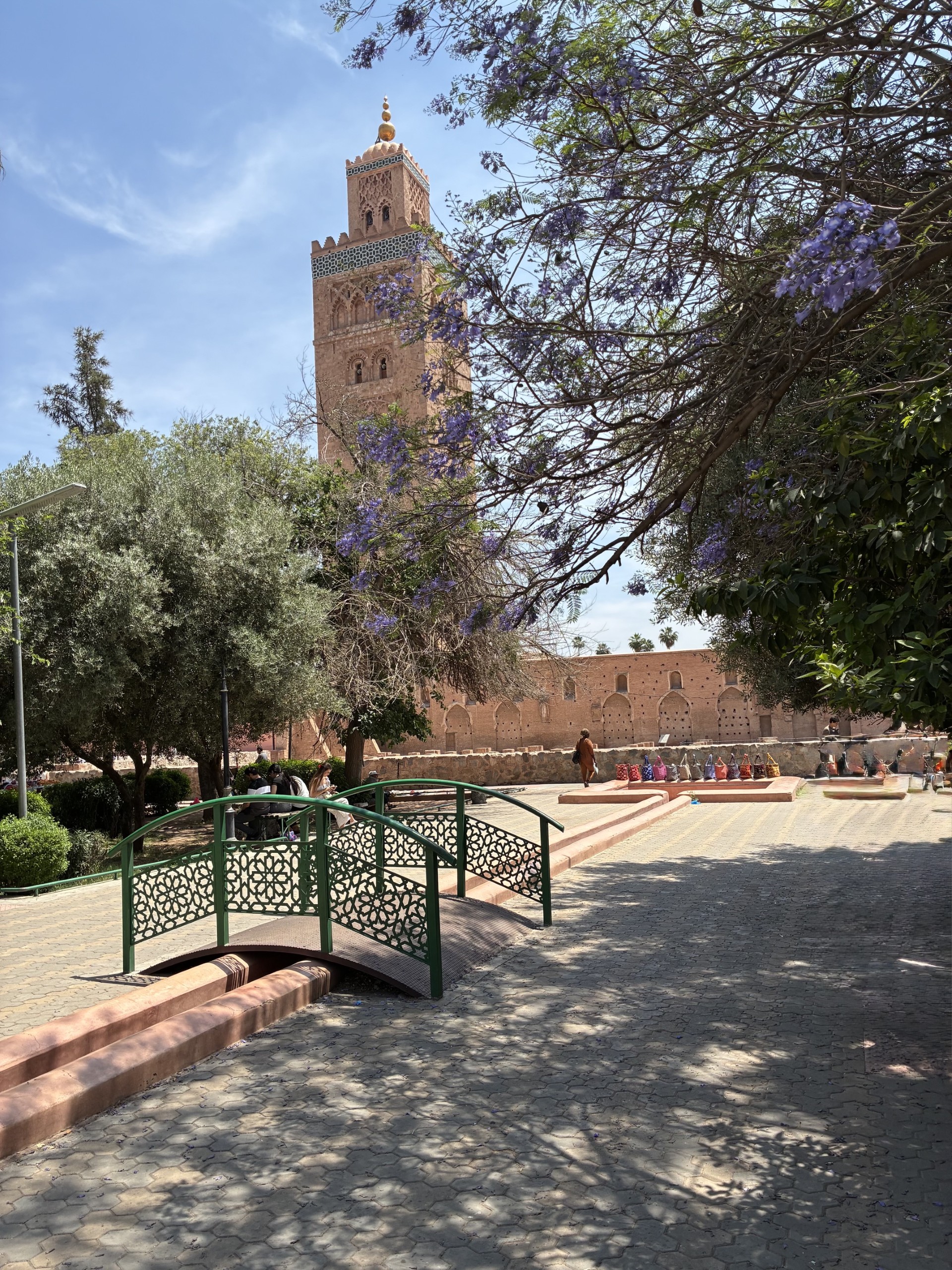 The Koutoubia Mosque's minaret rises over a Marrakech park, framed by a blooming jacaranda tree, with a decorative footbridge in the foreground and the mosque's ruined foundation walls behind it.