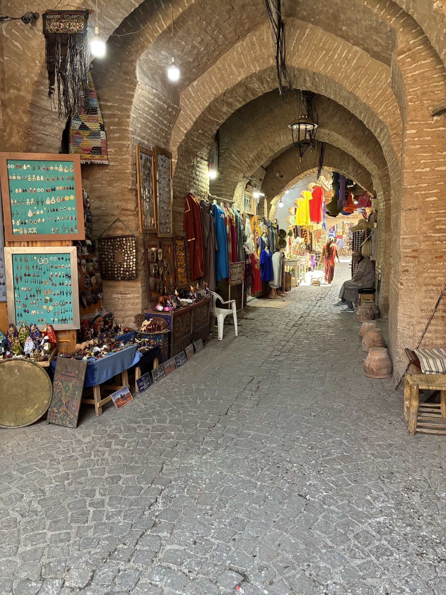Inside the covered souk of Marrakech, Morocco, a vaulted brick archway leads past stalls selling jewelry, djellabas, masks, and pottery, with a merchant seated near the cobblestone walkway.