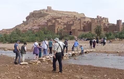 A group of travelers crosses a rocky riverbed on stones and sandbags toward Ait Ben Haddou, the fortified clay kasbah near Ouarzazate, Morocco, famous as a filming location for movies like Gladiator and Lawrence of Arabia.