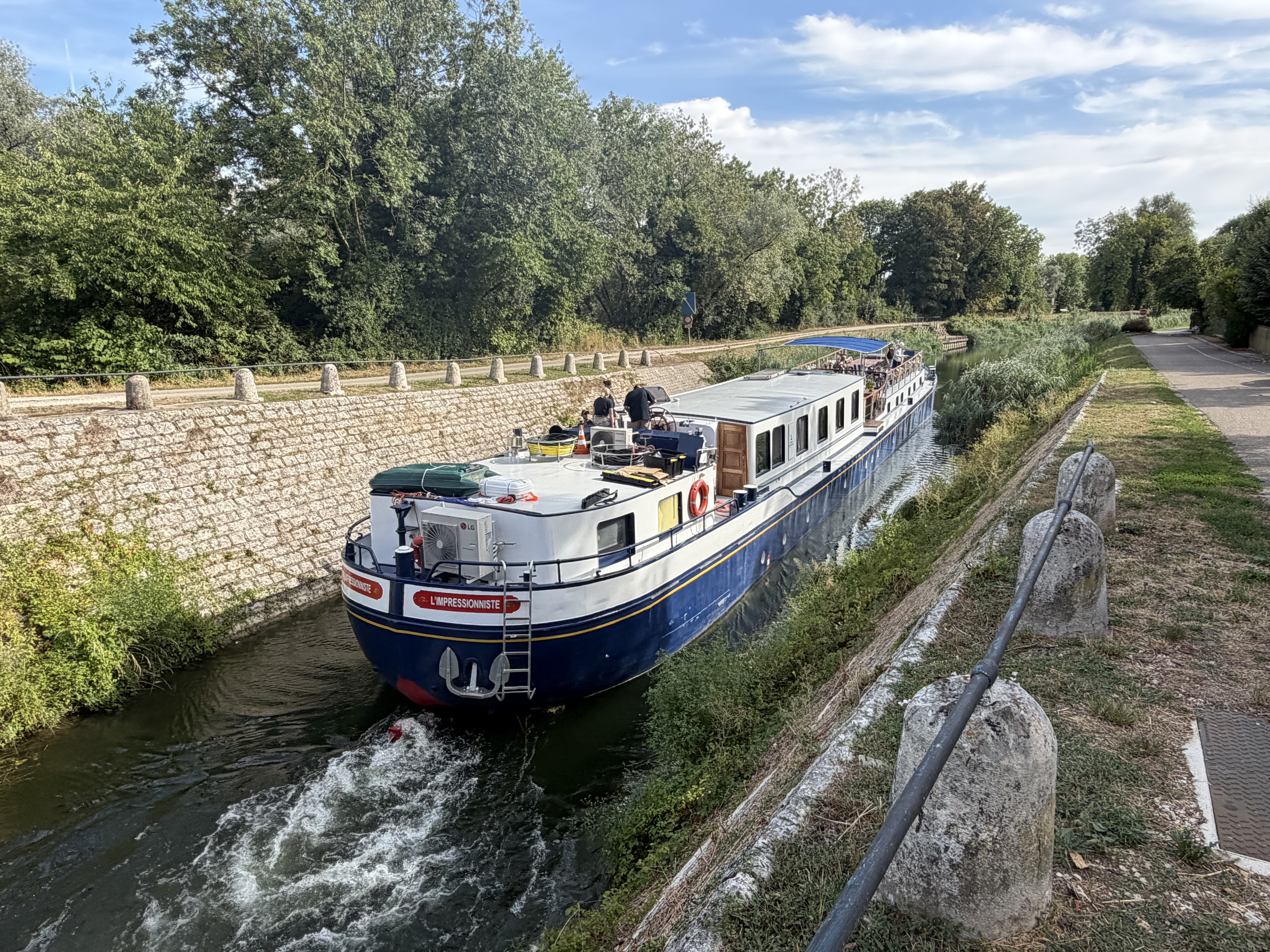 The hotel barge L'Impressionniste pushes through a narrow, stone-walled canal in rural France, crew at the helm on the upper deck.