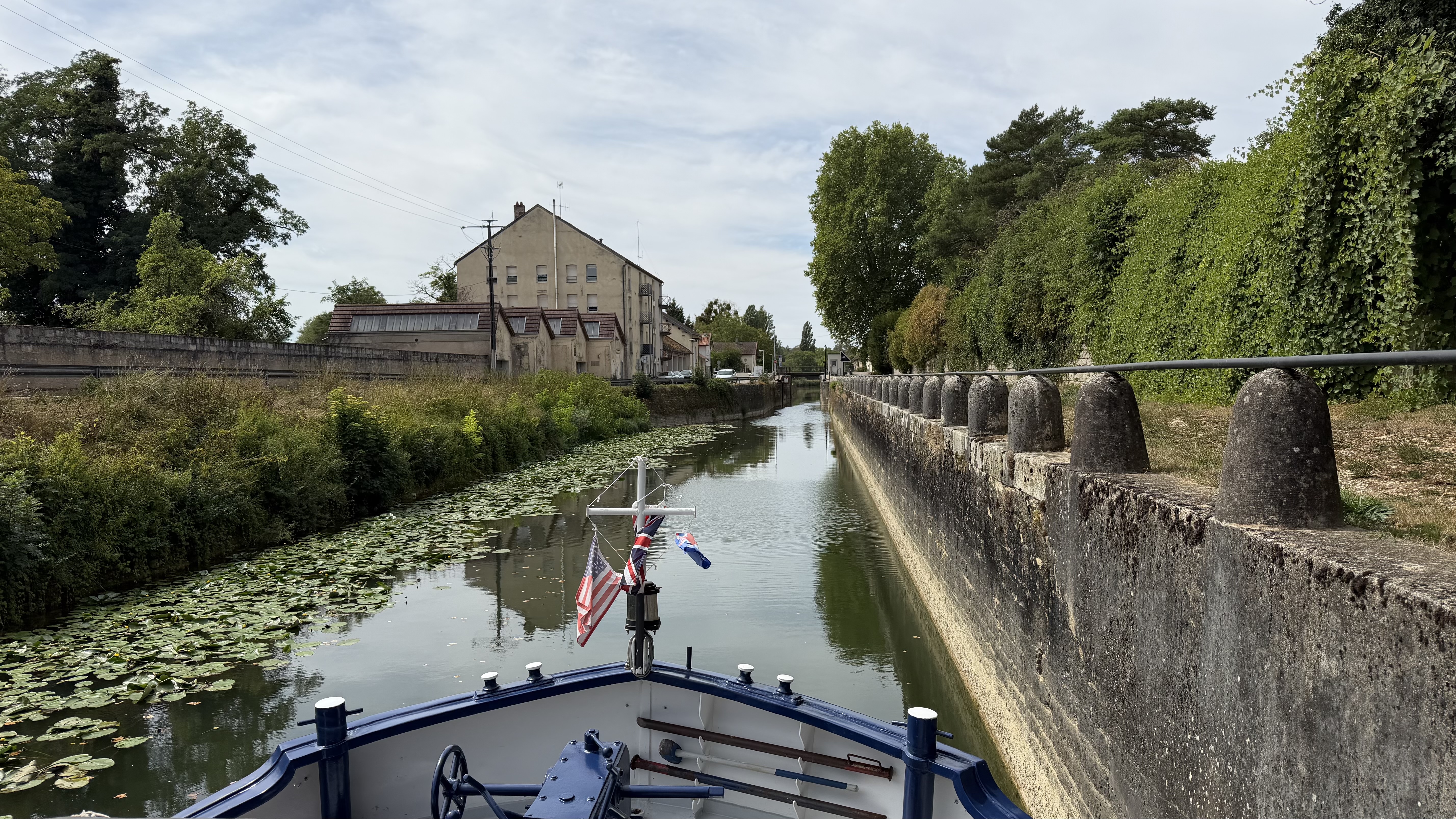 The bow of a hotel barge flying American, British, and French flags glides through a narrow, lily-covered canal in France, past a stone retaining wall and an old industrial building.
