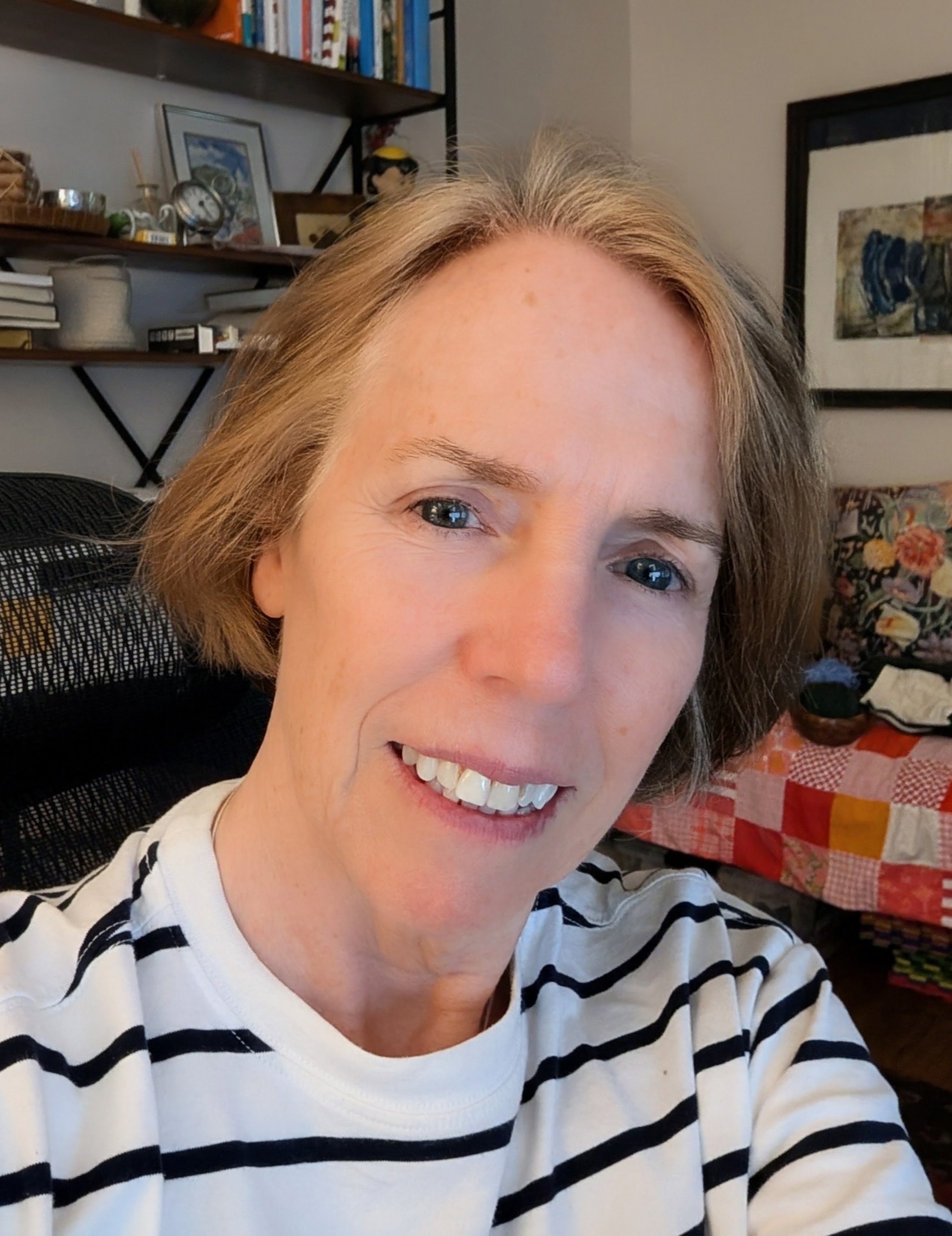 Justine smiling at the camera, with shoulder length blond hair and a white and navy striped top, in a home office with bookshelves.