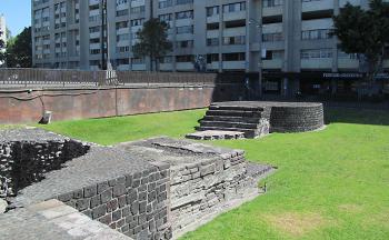 A view of the Tlatelolco archaeological zone showing a remnant of an Aztec staircase and temple base. In the background is one of the buildings of the Nonoalco complex.